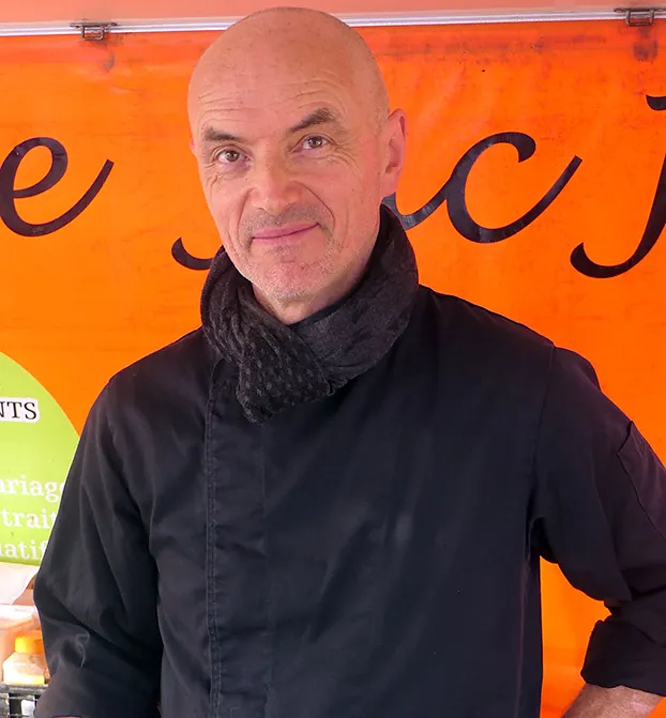 Jacky Gobbé, cuisinier, en veste noire et écharpe grise, souriant devant son stand de marché à la bâche orange.