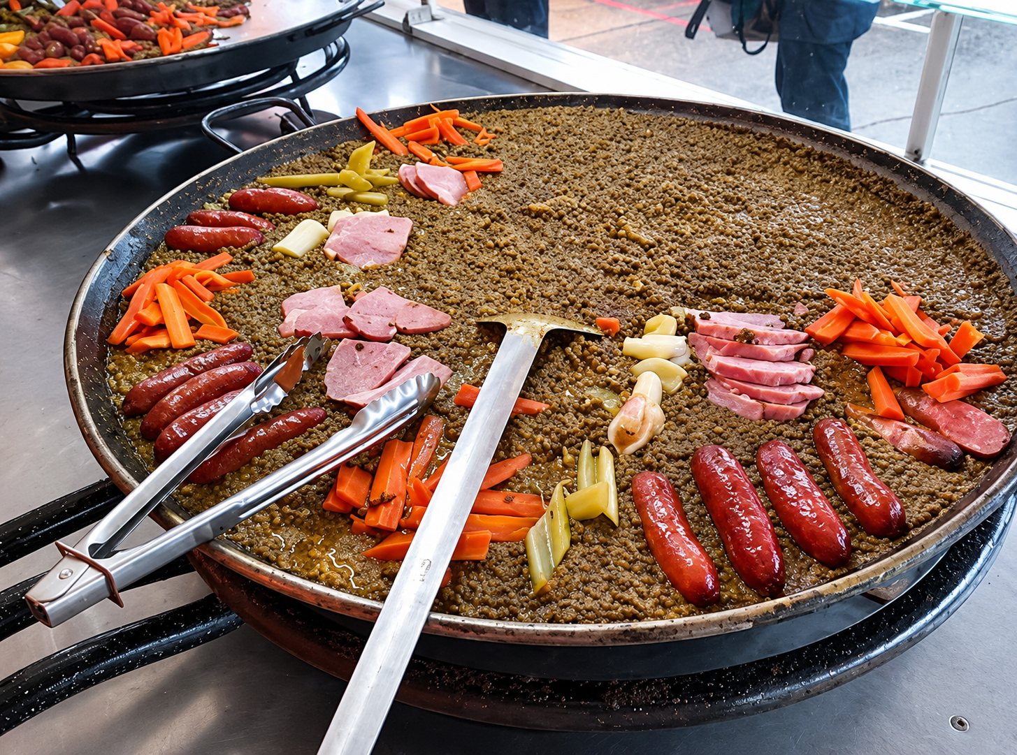 Une très grande poêle de paella en cours de préparation, garnie de saucisses, de bâtonnets de carotte, de tranches de jambon et de pâtes, avec une spatule et une pince en métal.