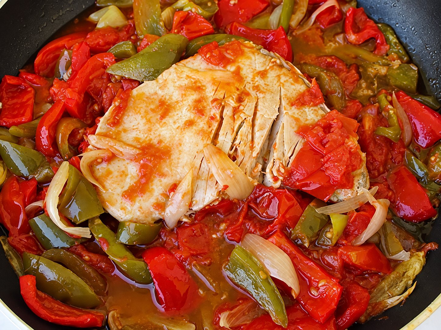 Une poêle contient des poivrons rouges et verts mijotés avec des oignons et un morceau de poisson nappé de sauce tomate.