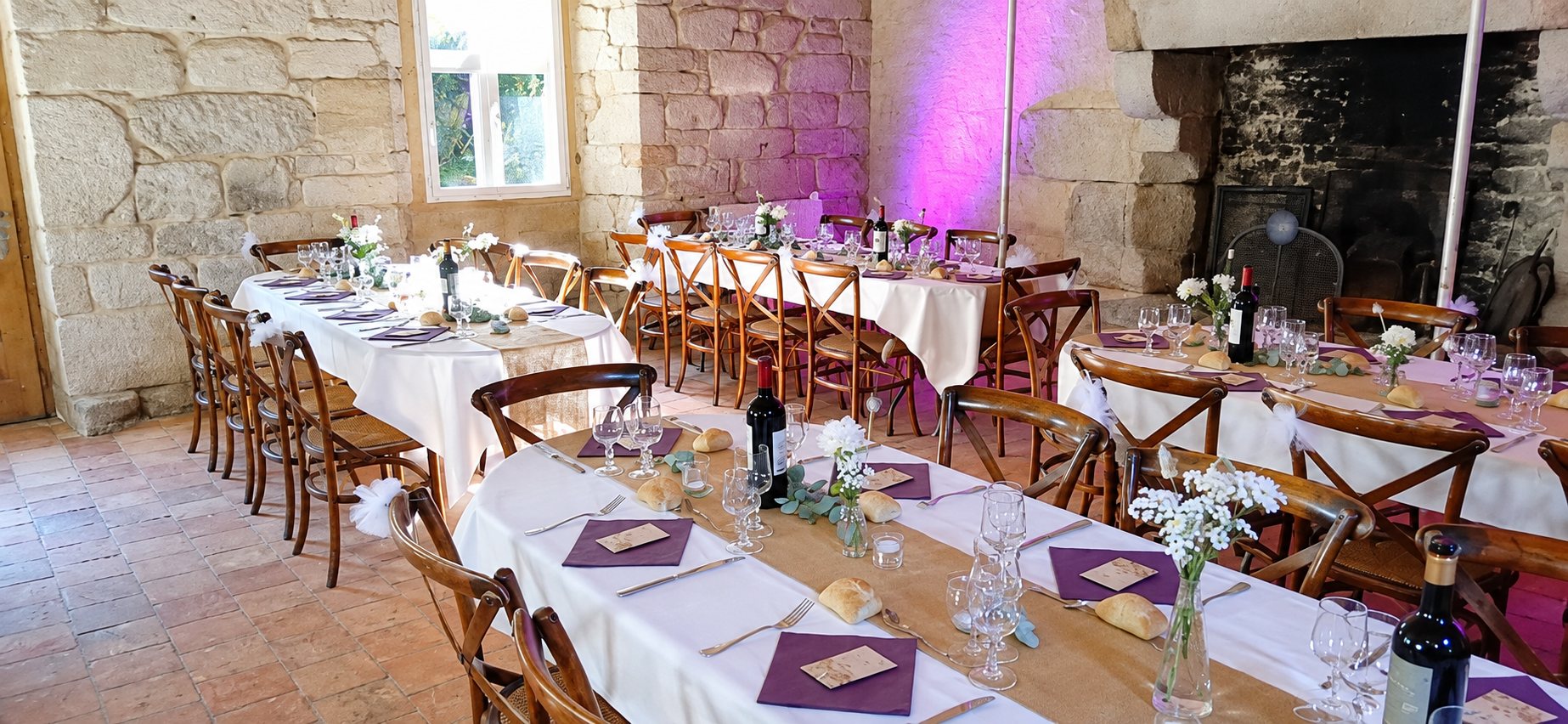 Une salle en pierres apparentes avec de longues tables blanches dressées, chaises en bois, verres, bouteilles de vin et petits bouquets de fleurs, sous une lumière violette.