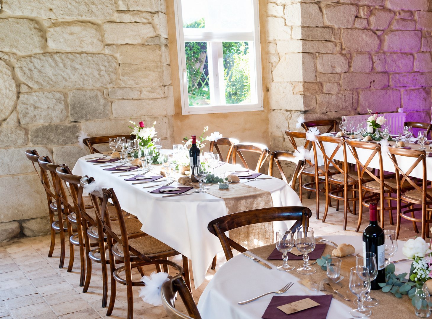 Des longues tables recouvertes de nappes blanches, dressées avec verres, assiettes et fleurs, dans une salle aux murs en pierre apparente éclairée par une fenêtre.